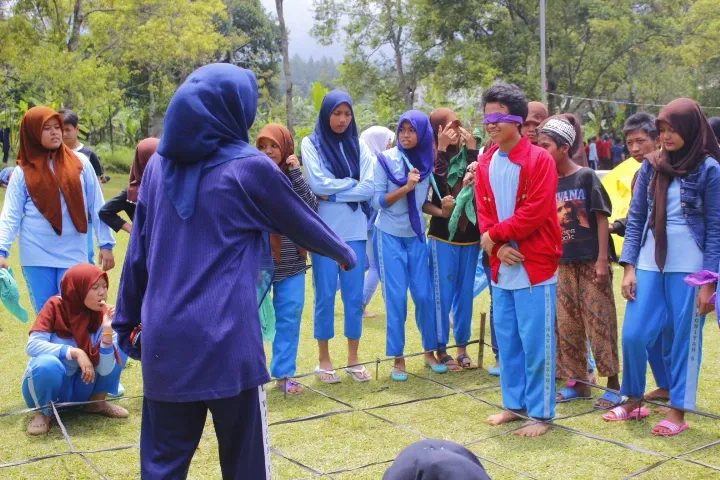 Siswa mengikuti permainan kelompok di lapangan rumput saat kegiatan luar kelas, salah satu siswa memakai penutup mata